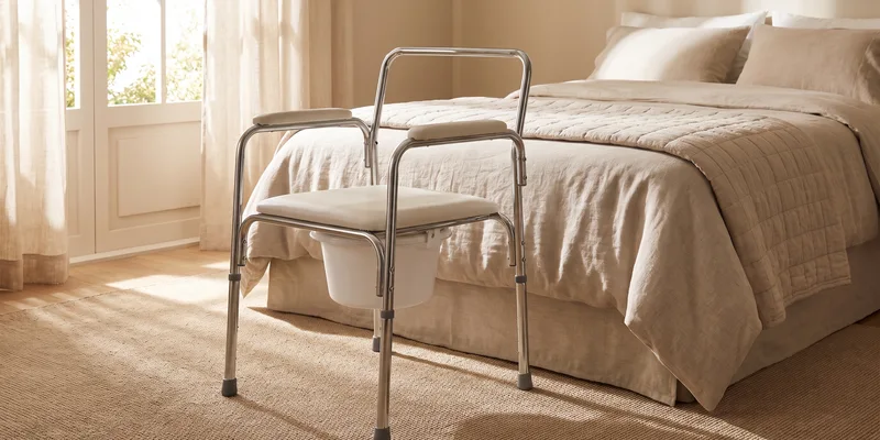 A steel framed commode chair with a removable bucket beneath the seat standing on the carpet beside a neatly made bed in a sunlit bedroom.