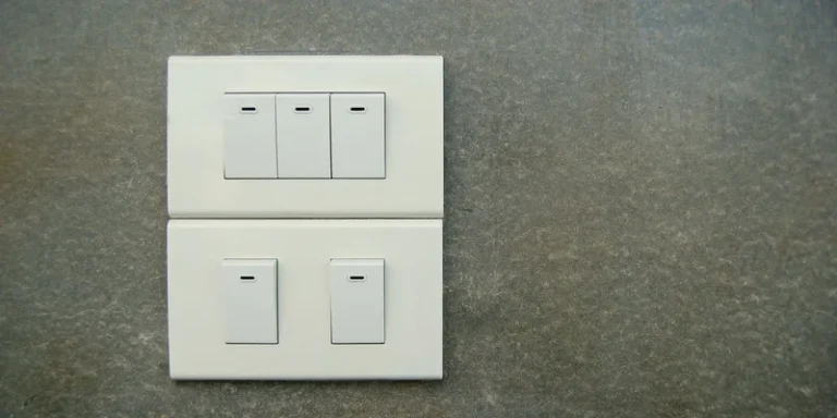 A single white rocker light switch on a pale painted wall beside an interior doorway in a sunlit hallway.