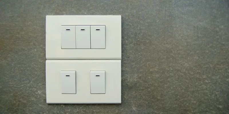 A single white rocker light switch on a pale painted wall beside an interior doorway in a sunlit hallway.