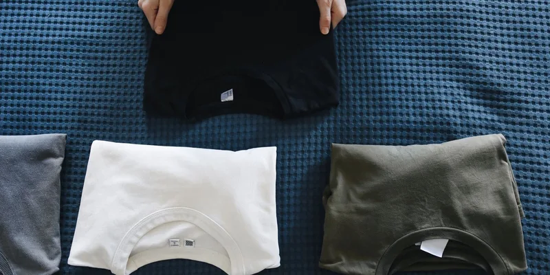A folded cardigan with magnetic front closures and a pair of side zip trousers laid out on a bedroom bench.