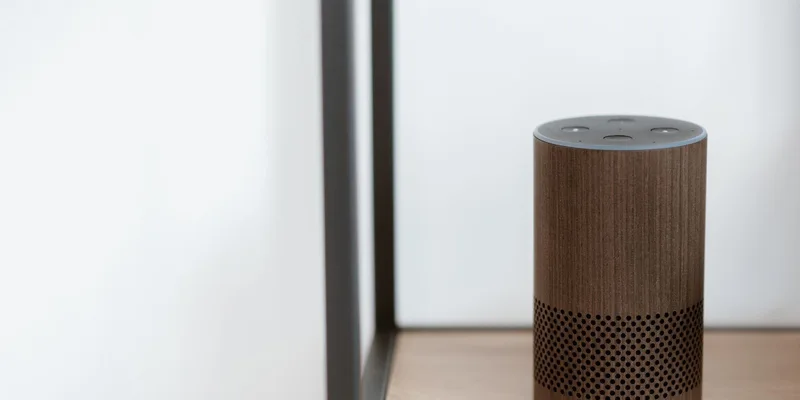 A small fabric covered smart speaker on a wooden dresser beside a table lamp in a warm living room.