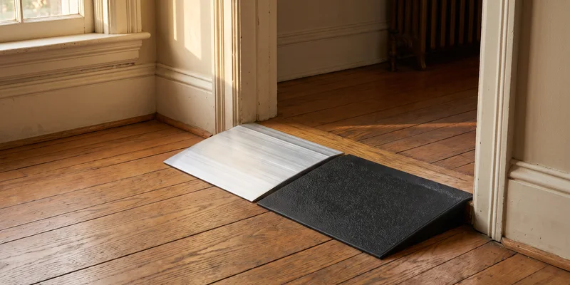 An aluminum threshold ramp and a black rubber threshold ramp resting side by side on a wooden floor at an interior doorway.