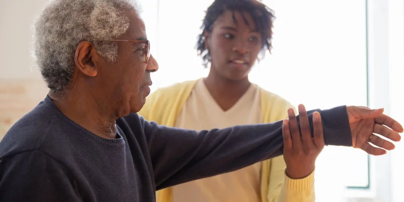 A caregiver supports an older man's arm during an exercise.