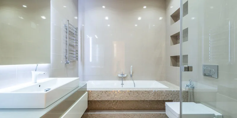 A shower chair, handheld shower head, long handled sponge and wall grab bar together in a bright tiled bathroom.