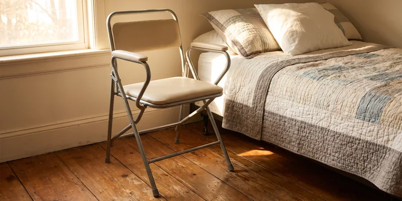 A folding steel bedside commode with padded arms standing beside a bed on a wooden floor.