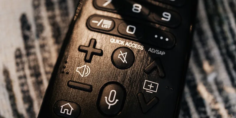 A simplified television remote with a few large keys resting on a sofa arm facing a living room television.