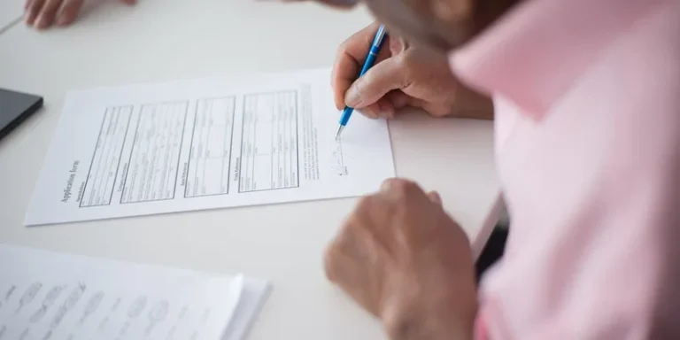 A hand fills in a paper form on a desk.