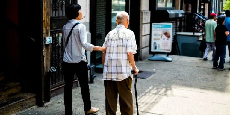 An older man with a cane is supported by a companion while walking.