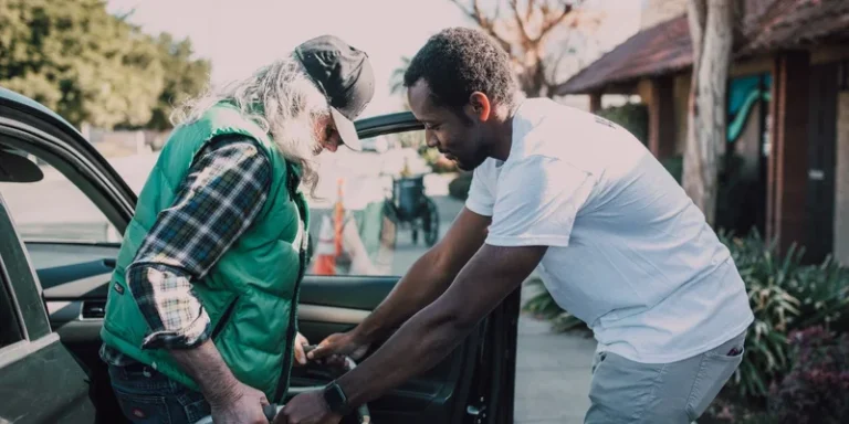A helper assists a seated older man beside a car.