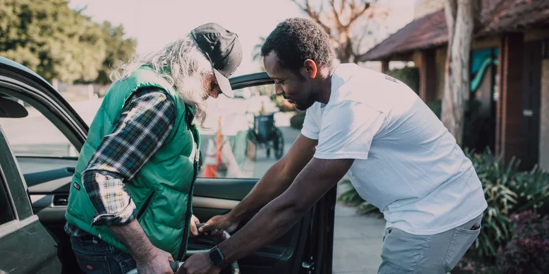 A helper assists a seated older man beside a car.