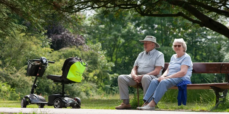 An older couple rest on a bench next to a mobility scooter.