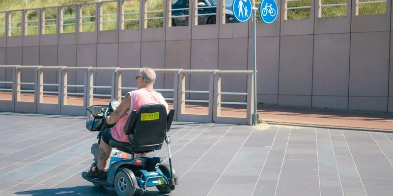 A senior rides a four-wheel mobility scooter along a wide paved walkway.