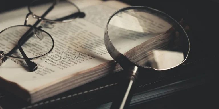 A lighted handheld magnifier and a stand magnifier on a desk beside a lamp.