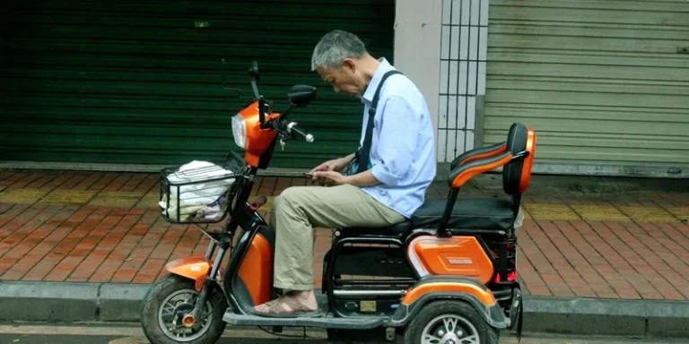 An older man seated on a three-wheel mobility scooter on the sidewalk.