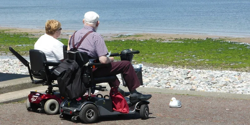 An older couple seated on mobility scooters beside the water.