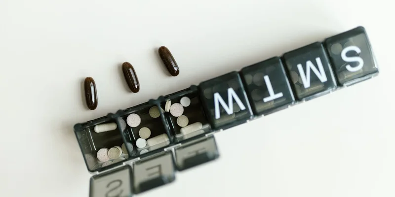A weekly pill organizer with its lids open beside a monthly tray on a kitchen counter.