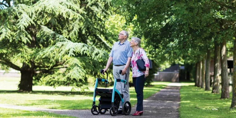 An older couple walk together with a rollator on a park path.