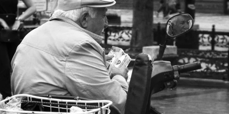 A senior man sits reading on a mobility scooter in a park.