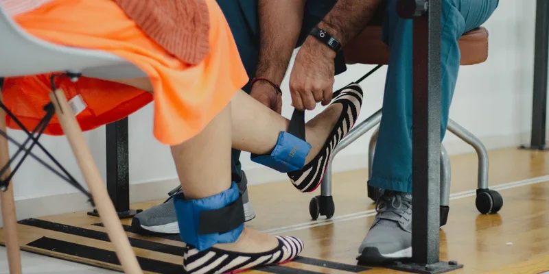 A therapist works with a seated patient wearing ankle weights.
