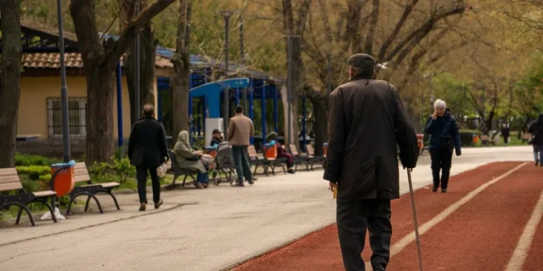 An older man walks with a cane along a tree-lined park path.